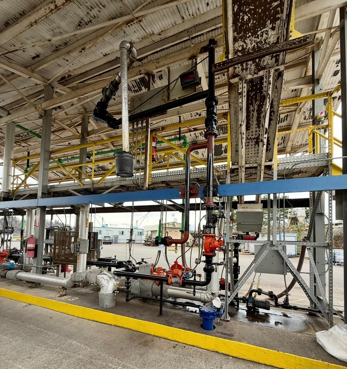 Industrial manufacturing facility interior with metal scaffolding, machinery, and blue platform structure under metal ceiling with overhead piping and equipment